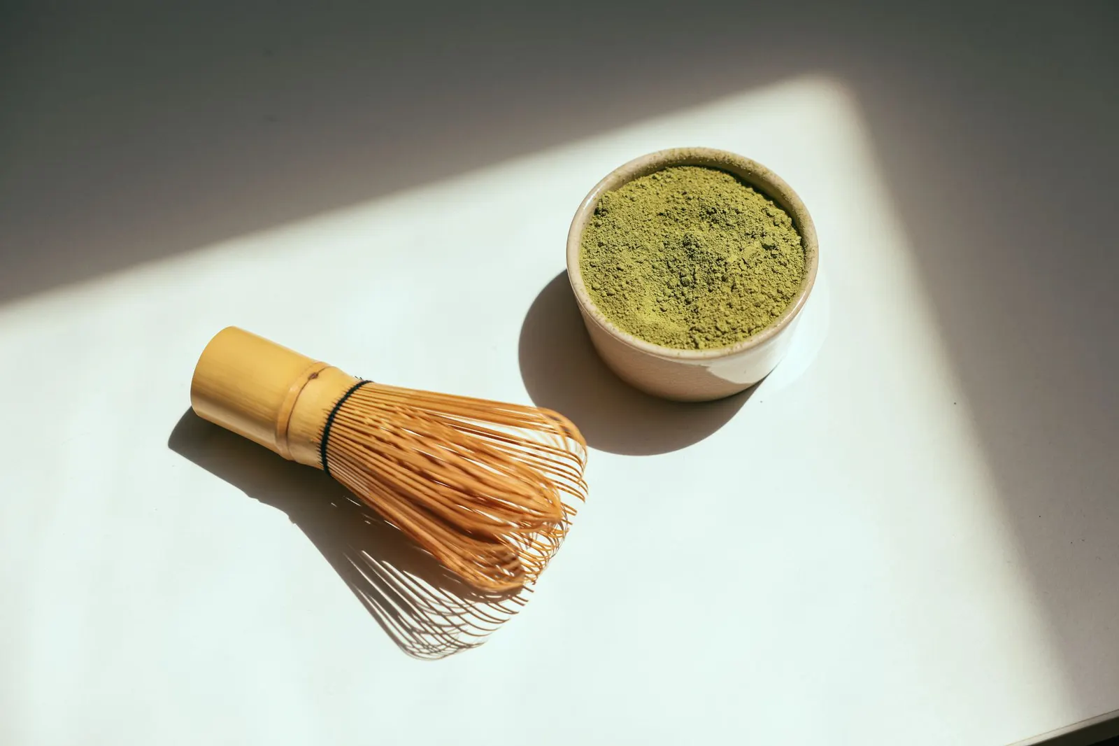 Matcha powder in a bowl beside a bamboo whisk in bright sunlight